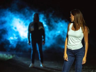 Woman in white tank top looks back at a shadowy figure in blue mist at night.