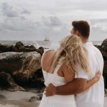 Couple embracing on rocky beach with sailboat in the distance.