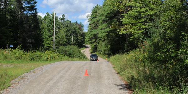 A black car parked on a gravel road with orange cones and surrounded by dense green trees.