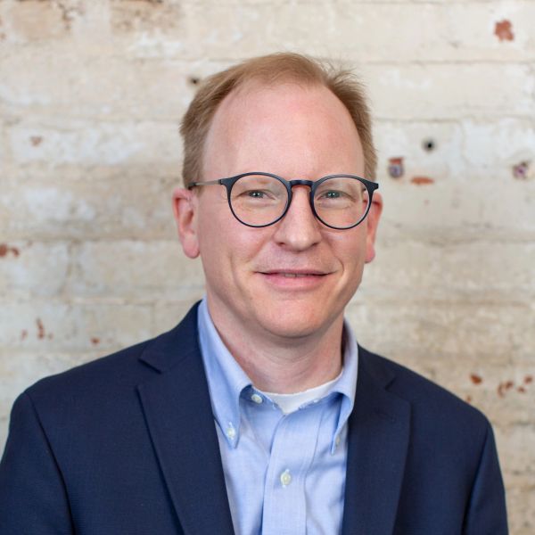 Man in glasses and suit smiling in front of a brick wall.
