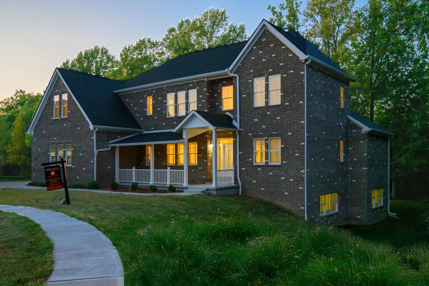 Large brick house with lights on and a for sale sign at dusk.