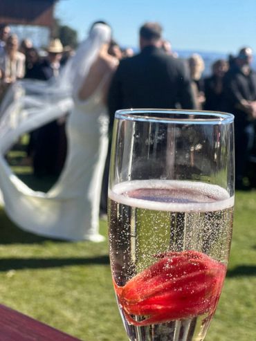 A glass of sparkling drink with a red flower, blurred wedding couple in background.