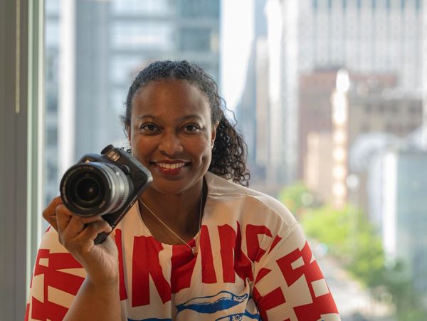 A smiling woman holding a camera, with a cityscape background.
