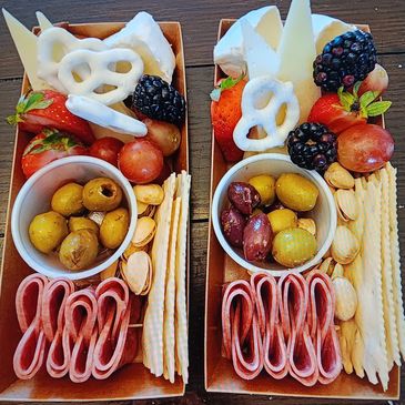 Two charcuterie boxes with fruits, cheese, crackers, olives, and nuts.