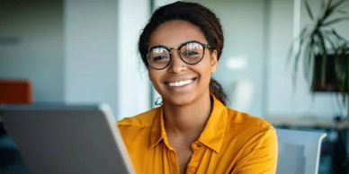 Smiling woman in glasses working on a laptop in a bright office.