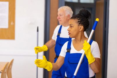Two janitors in blue overalls and yellow gloves cleaning a room.