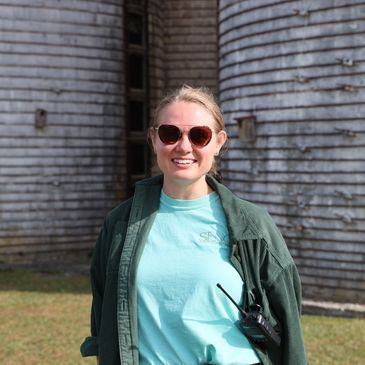 Smiling woman in sunglasses and casual clothing stands outdoors near weathered wooden structure.