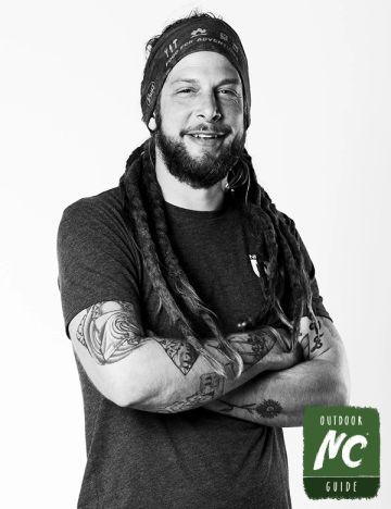 Man with dreadlocks and tattoos smiling, wearing a bandana and dark t-shirt.