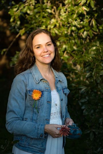 Smiling woman in denim jacket with flowers, standing outdoors in sunlight.