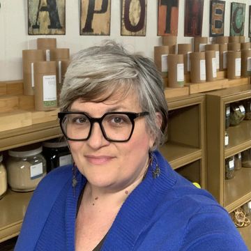 Smiling woman with glasses in a cozy apothecary setting.