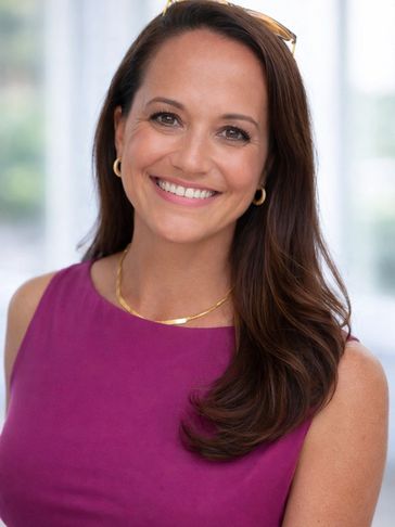 Smiling woman in a purple top with gold accessories and long brown hair.