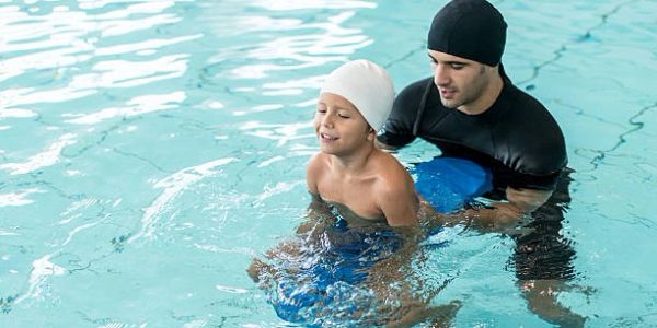 A man teaches a child to swim in a pool, both wearing swim caps.