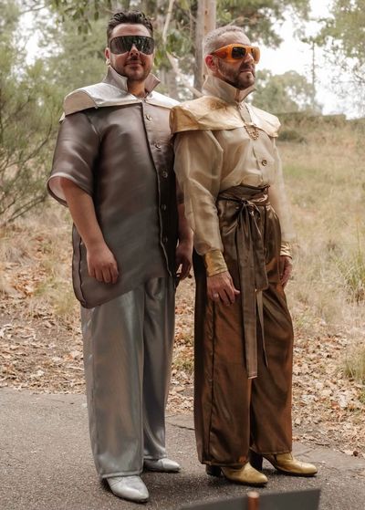 Two men in futuristic metallic outfits with sunglasses outdoors.