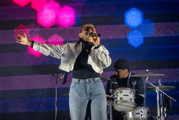 Female singer passionately performing on stage with drummer in the background.