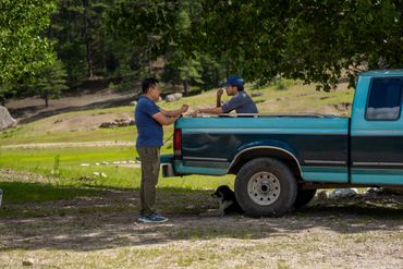 Two men sharing drinks by a pickup truck in a natural setting.