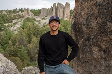 Smiling man in a black sweater and cap posing among rocky cliffs and greenery.