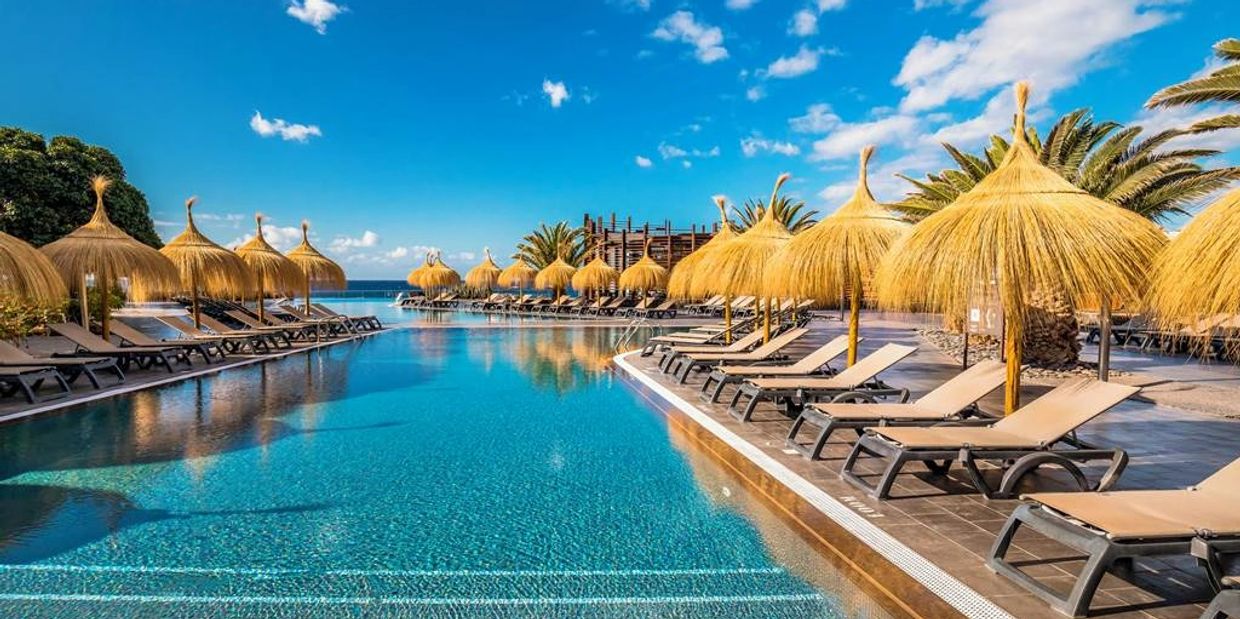 Sunny resort pool with thatched umbrellas and lounge chairs under a bright blue sky.