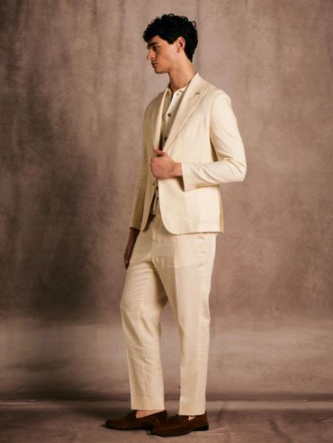 Man in cream-colored suit and brown shoes posing against a textured backdrop.