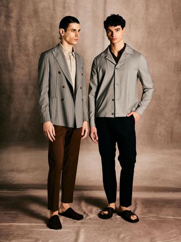 Two men modeling stylish, neutral-toned outfits against a textured backdrop.
