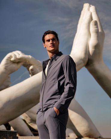 A man in gray outfit stands before giant praying hand sculptures under a blue sky.