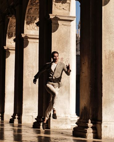 Man in stylish outfit walking between sunlit ancient columns.