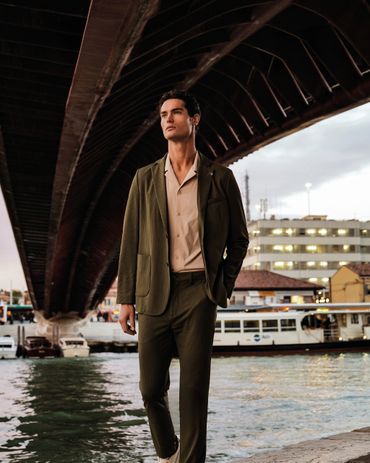 A man in a green suit walks confidently near a waterfront under a bridge.