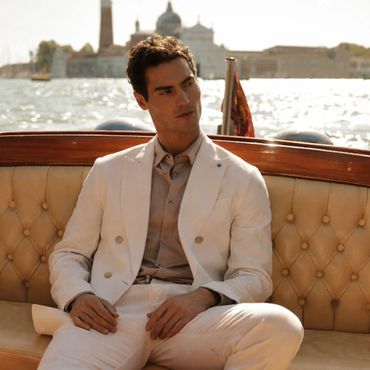A man in a white suit sitting on a boat with a scenic waterfront background.