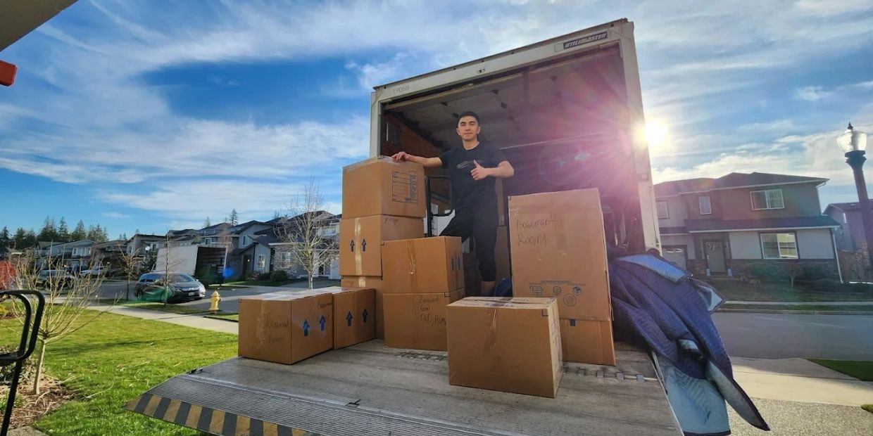 Man stands on moving truck ramp surrounded by cardboard boxes.