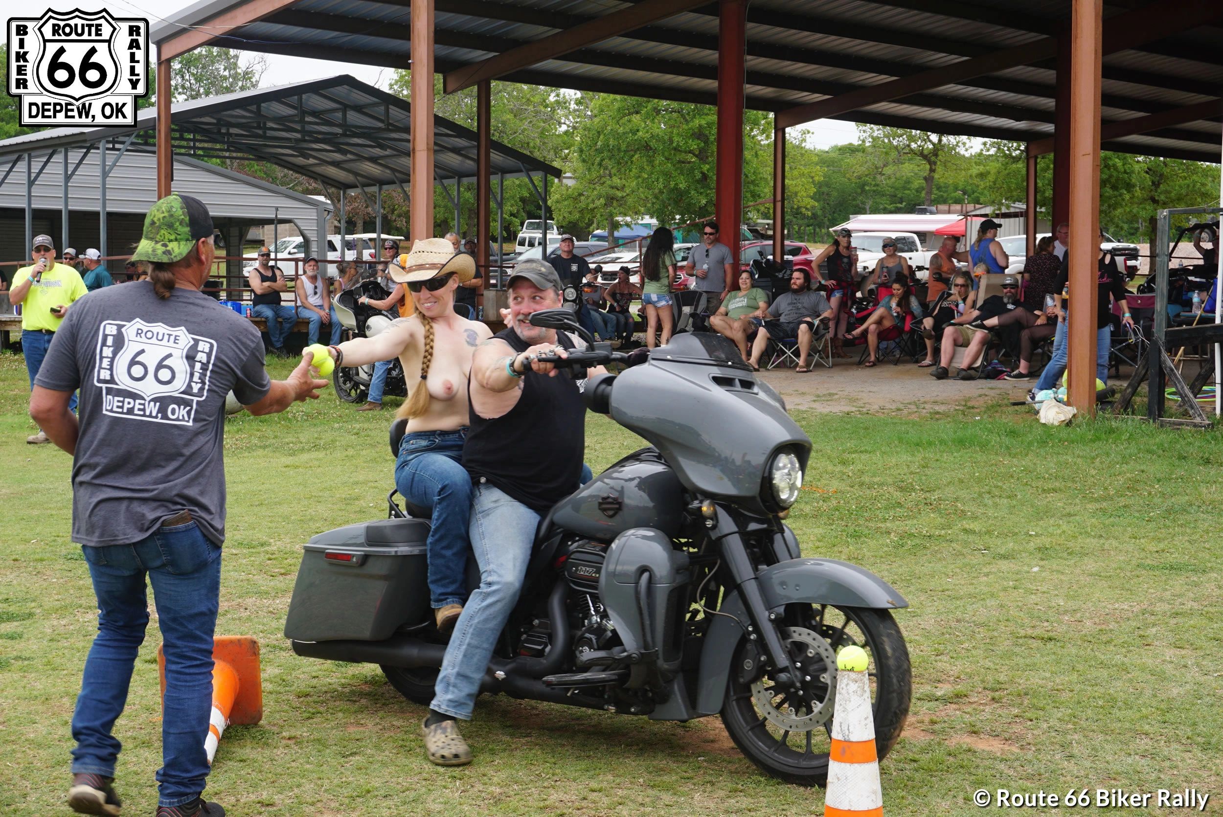 A topless woman on a motorcycle hands a ball to a man at a Route 66 Biker Rally.
