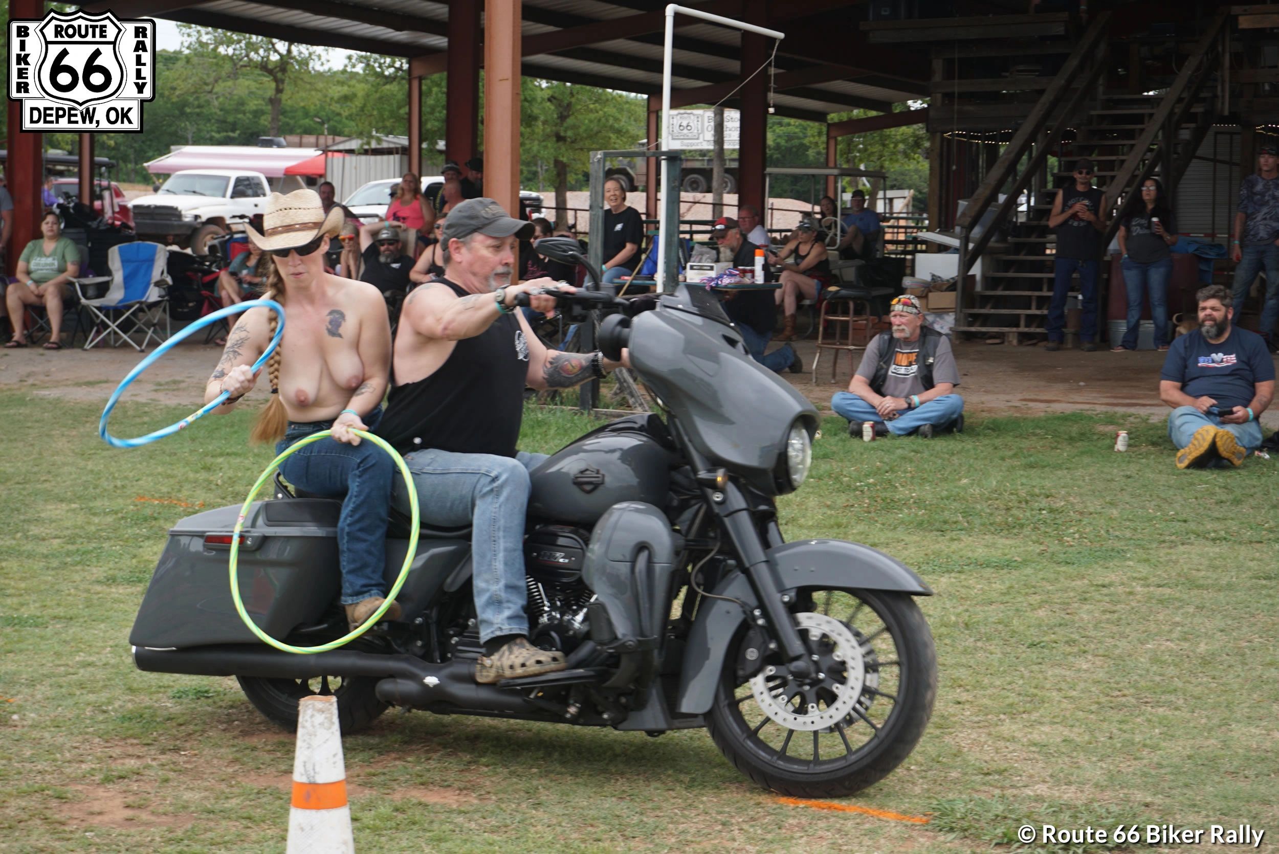 A topless woman and a man ride a motorcycle while holding hula hoops at a biker rally.