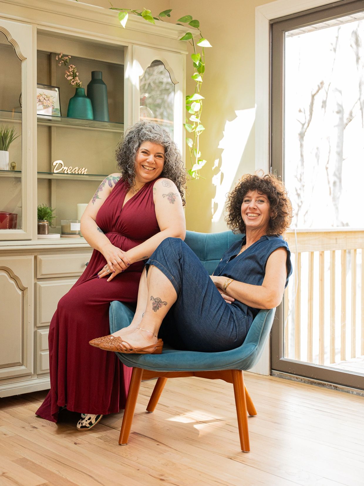 Two women with curly hair smiling indoors with tattoos visible.