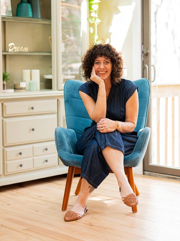 Woman with curly hair sitting on a blue chair, smiling warmly indoors.