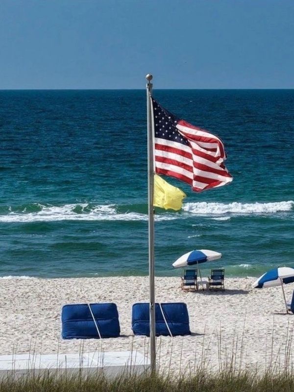 American flag and yellow flag flying on a beach with blue chairs and umbrellas.