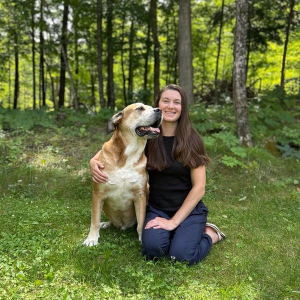 A woman happily kneeling in the grass with her large dog in a wooded area.