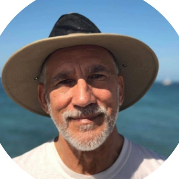 Smiling man wearing a wide-brimmed hat near the ocean on a sunny day.