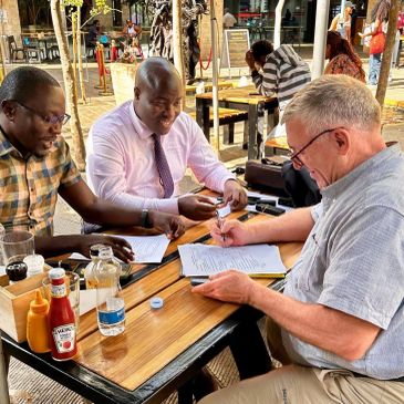 Three men reviewing and signing documents at an outdoor table.
