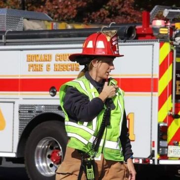 Firefighter in gear speaking into a radio near fire truck.