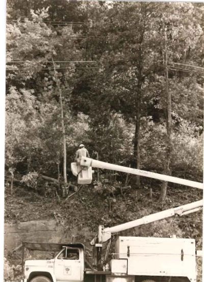 Utility worker in bucket truck repairing power lines by wooded hillside.