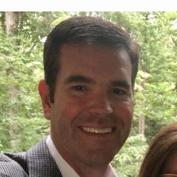Smiling man in a checkered jacket with a green leafy background.