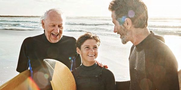 Three generations in wetsuits holding surfboards at the beach, smiling.
