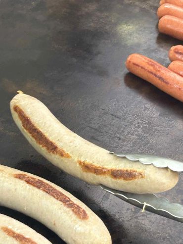 Grilled sausages cooking on a flat grill with tongs holding one sausage.