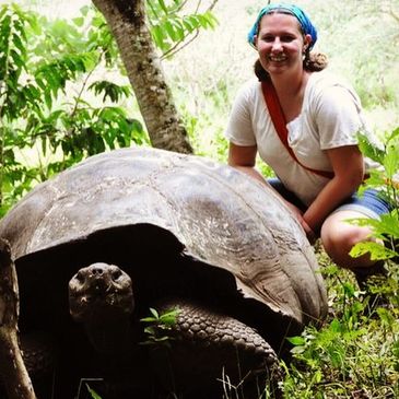 Woman posing with a giant tortoise in a lush outdoor setting.