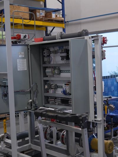 Open electrical control panel in an industrial setting with machinery and shelves.