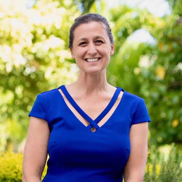 Smiling woman in a blue dress outdoors with greenery in the background.