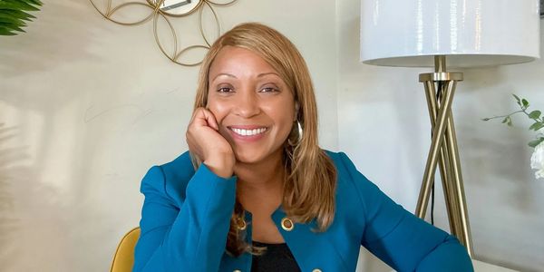 Smiling woman in a teal blazer sitting at a desk with a lamp and wall decor.