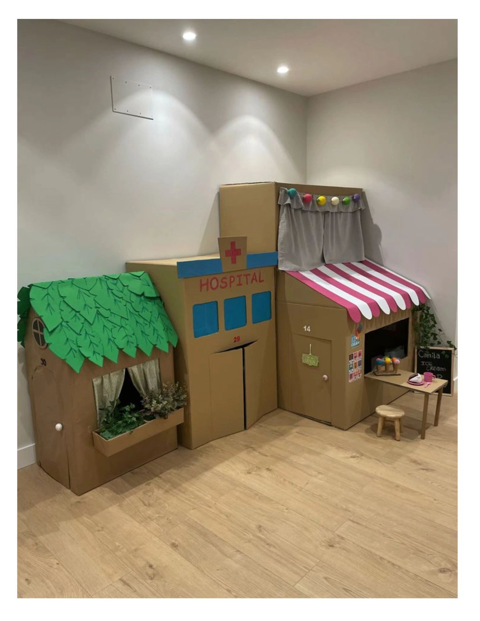 Cardboard playhouses designed as a hospital, a house, and an ice cream shop in a room.