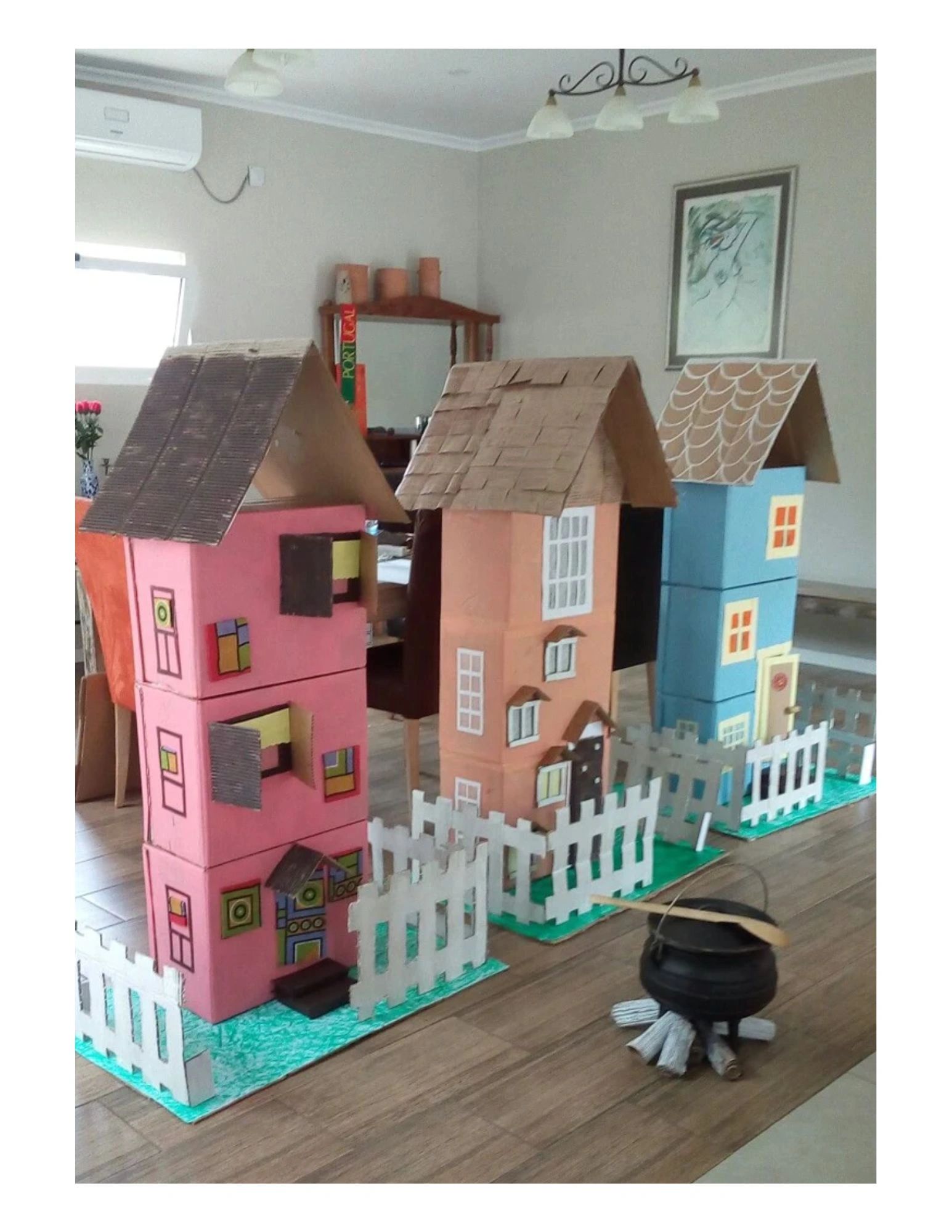 Three colorful cardboard model houses with fences and a small black cauldron on a wooden floor.
