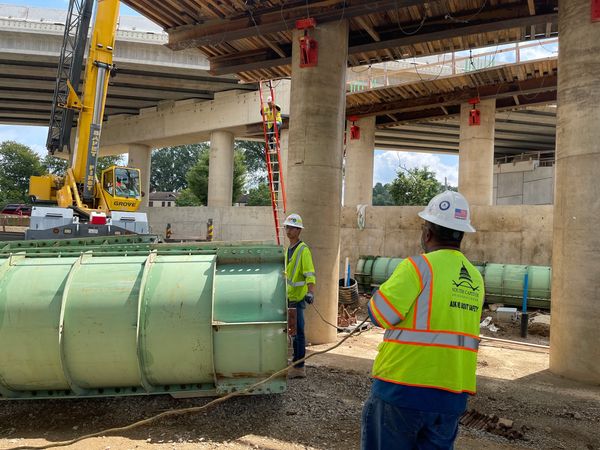 Construction workers inspecting large green piping at a bridge site.