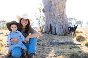 Woman and child wearing hats sit outdoors near a large tree with a dog in the background.