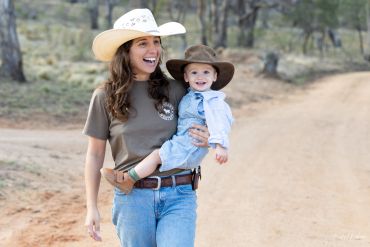 A smiling woman holding a baby, both wearing cowboy hats outdoors.
